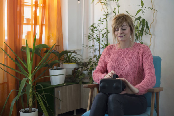 mujer en suéter rosa en casa disfrutando usando gafas de realidad virtual en sillón moderno Mirando a su alrededor y utilizando gestos con las manos
 - Foto, Imagen