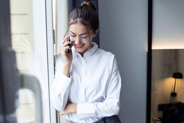 Shot of pretty young businesswoman talking with her smartphone standing near the window at hotel room. - Fotoğraf, Görsel