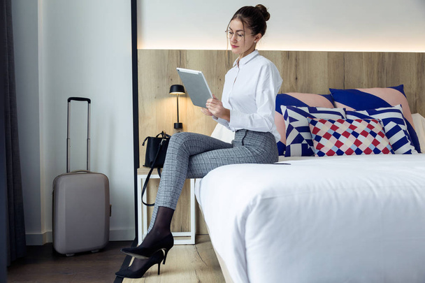 Shot of attractive young businesswoman using her digital tablet sitting on the bed at hotel room. - Valokuva, kuva