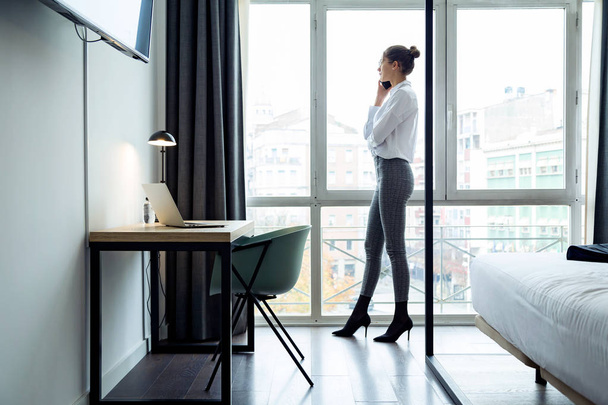Shot of stylish young businesswoman talking with her smartphone and looking sideways standing near the window at hotel room. - Фото, изображение
