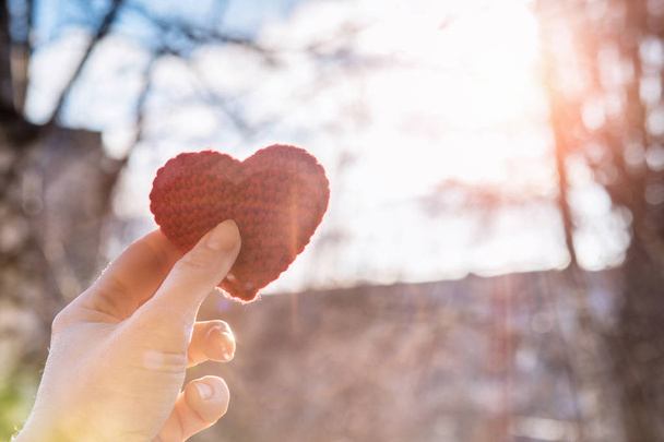 Una mano femminile tiene un cuore a maglia rosso vivo sullo sfondo del paesaggio urbano dell'alba del mattino. Il concetto di relazioni familiari
. - Foto, immagini