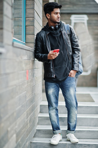 Κομψό και casual Ασίας άνθρωπος σε μαύρο δερμάτινο μπουφάν, ακουστικά με κόκκινο κινητό τηλέφωνο στα χέρια που τίθενται στον δρόμο. - Φωτογραφία, εικόνα