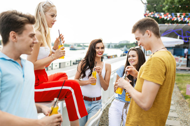 A company of good-looking friends laughing and drinking yellow cocktails in the nice cafe next to the river. Cheers. Entertainment, having good time. River is in the background. - Фото, зображення