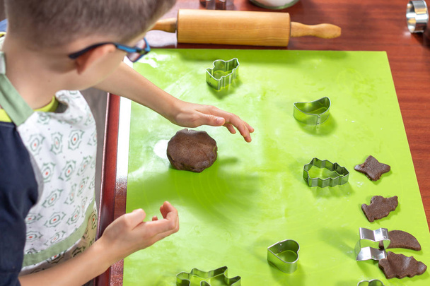 Kleine Köchin. Der Junge bereitet Zimtkuchen für Plätzchen zu. Kinderhand formt einen Kuchen auf einem Silikontisch. Metallformen und eine Holzwalze liegen daneben. - Foto, Bild