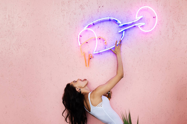 The girl poses on the pink wall, pretending to drink from a glass made of neon tubes attached to the wall - Foto, afbeelding