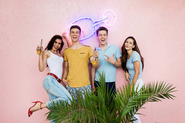 A company of good-looking friends laughing, drinking yellow cocktails is standing in front of the pink wall and behind a green plant in the nice summer cafe. Cheers. Entertainment, having good time - Foto, Bild