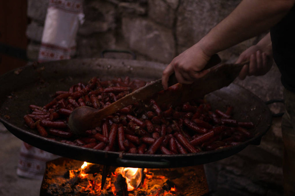 Cocinero tradicional en una sartén de hierro fundido. Salchichas asadas sobre una llama abierta. Cocinar para mucha gente. Cocina al aire libre en las montañas. Fuego.
. - Foto, Imagen