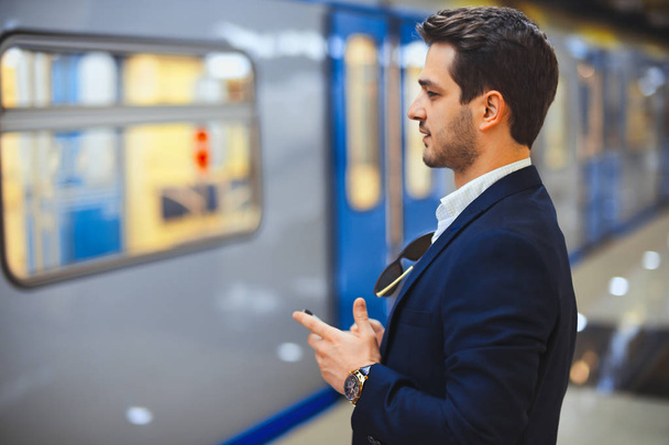 Young attractive businessman texting message in mobile phone while waiting for train in metro. Enterpreneur solving working issues. Lating on work. - Φωτογραφία, εικόνα