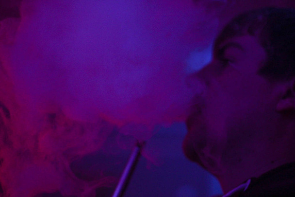 Πορτραίτο ενός ανθρώπου που καπνίζει ένα ναργιλέ και εκπνέετε καπνό με μια μωβ μαρτυρία στο παρασκήνιο. Cafe. Μπαρ. Καλλιτεχνικές φωτογραφίες - Φωτογραφία, εικόνα