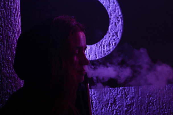 Portret van een meisje die een waterpijp en uitademen rook met een paarse getuigenis in de achtergrond rookt. Cafe. Bar. Artistieke foto 's - Foto, afbeelding