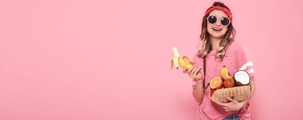 Beautiful young girl in pink t-shirt and glasses, holding sexy bananas and full bag of fruit on pink background. Summer and Rest. Place for text. Advertising concept - Fotografie, Obrázek