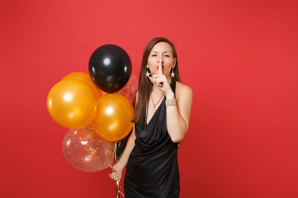 hübsche Frau in schwarzem Kleid, die mit dem Finger auf den Lippen schweigt, shhh Geste feiert und Luftballons isoliert auf rotem Hintergrund hält. Frohes neues Jahr Geburtstag Attrappe Urlaub Party-Konzept - Foto, Bild