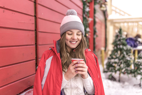 Girl red winter jacket stands in wooden shutters. In his hand holds cup of hot coffee and tea, warms. Concept of outdoor recreation resort, happy woman smiling with eyes closed fantasize and dream. - Photo, Image
