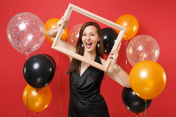 Mujer joven alegre en pequeño vestido negro celebrando y sosteniendo marco de imagen en globos de aire de fondo rojo brillante. Día Internacional de la Mujer, Feliz Año Nuevo concepto de fiesta simulada de cumpleaños
 - Foto, Imagen