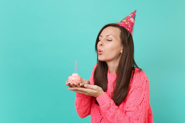 Stunning young girl in knitted pink sweater birthday hat with closed eyes blowing out candle on cake in hands isolated on blue background studio portrait. People lifestyle concept. Mock up copy space - Photo, Image