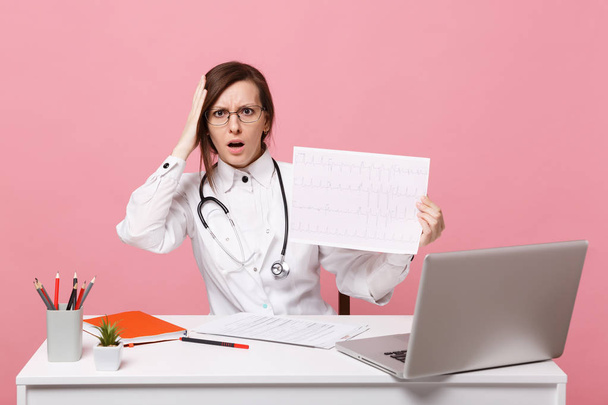 Female doctor sits at desk works on computer with medical document heart ekg in hospital isolated on pastel pink wall background. Woman in medical gown stethoscope. Healthcare medicine concept - Photo, Image
