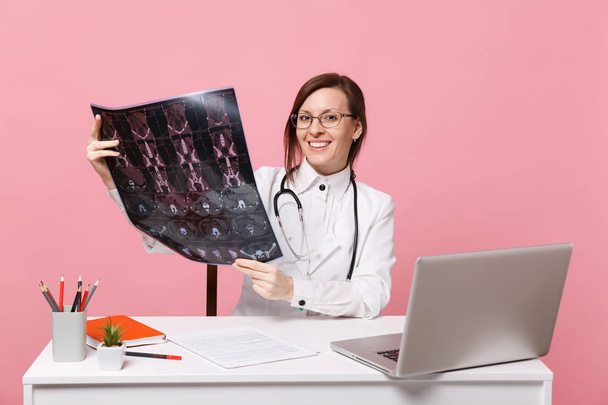 Naislääkäri istuu työpöydän ääressä tietokoneella, jossa on lääketieteellinen asiakirja pidä röntgenkuva sairaalassa eristettynä pastellin vaaleanpunaiselle seinälle. Nainen lääkärinpuvussa, stetoskooppi. Terveydenhuollon lääketieteellinen käsite
 - Valokuva, kuva