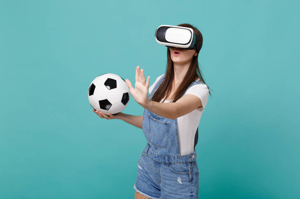 Удивительная девушка футбольный болельщик смотрит в наушники, держа футбольный мяч играть прикоснуться что-то вроде нажмите на кнопку, указывая на плавающий виртуальный экран изолированы на синем бирюзовом фоне стены
 - Фото, изображение