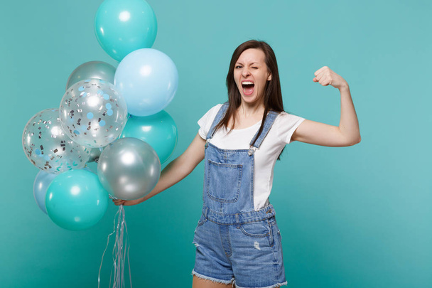 Loco divertido gritando mujer joven parpadeando, mostrando bíceps, músculos celebrando, sosteniendo globos de aire de colores aislados sobre fondo azul turquesa. Fiesta de cumpleaños, gente concepto de emociones
 - Foto, imagen
