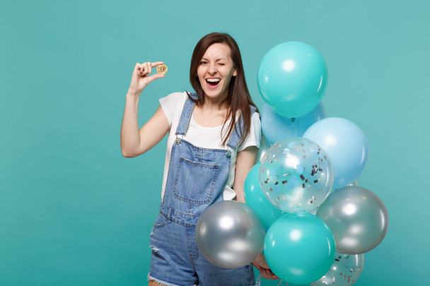 Wesoły migający młoda kobieta gospodarstwa bitcoin, walutową i świętuje z kolorowych balonów na białym tle na tle niebieski turkus ściany. Urodziny wakacje, koncepcja emocje ludzi - Zdjęcie, obraz
