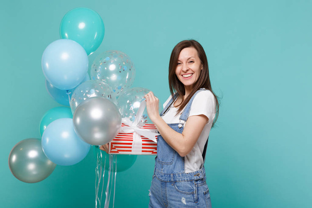 Pretty young woman hold red striped present box with gift ribbon, celebrating with colorful air balloons isolated on blue turquoise background. Valentine's Day, Women's Day, birthday, holiday concept - Photo, Image