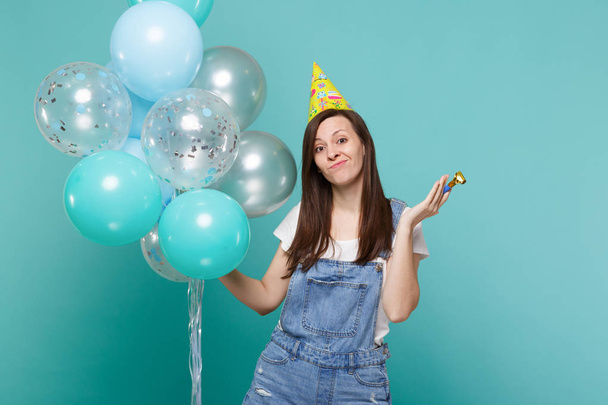 Saai verbijsterd jonge vrouw in denim kleding, verjaardag hoed bedrijf pijp vieren met kleurrijke lucht ballonnen geïsoleerd op blauwe turkooizen achtergrond. Vakantie verjaardagsfeestje, mensen emoties concept - Foto, afbeelding