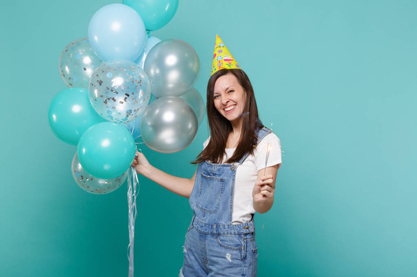 Attractive smiling young woman in birthday hat holding burning sparkler, celebrating with colorful air balloons isolated on blue turquoise background. Birthday holiday party, people emotions concept - Foto, imagen