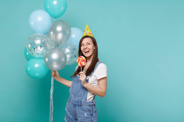 Thinkful young woman in birthday hat looking up, holding round lollipop, celebrating with colorful air balloons isolated on blue turquoise background. Birthday holiday party, people emotions concept - Foto, imagen