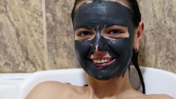 Woman with black cleansing cream mask on face looking at camera and smile. Skin care concept - Footage, Video