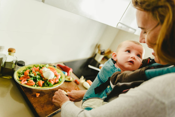 Bebê sorridente portado em mochila transportadora de bebê olhando para sua mãe enquanto ela cozinha, conceito de conciliação familiar
 - Foto, Imagem