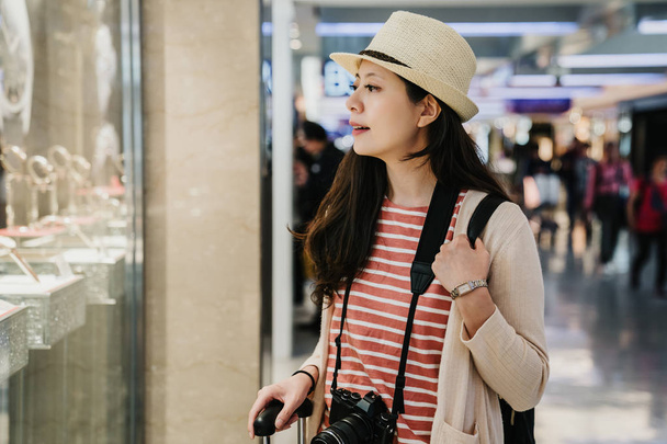 asian female travel photographer standing outside showing window of watch jewelry shop in duty free zone. chinese young girl tourist in hat window shopping in tax free area finding buying present. - Foto, immagini