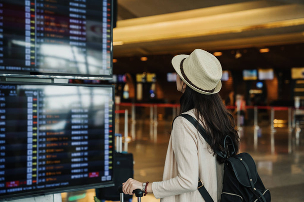 Hátulnézet fiatal utazók turisztikai nő keres ütemterv ütemterv a lobbi csarnokában nemzetközi repülőtér várja a kalapban. női utas utazik külföldön a menekülés hétvégén. Légi járat koncepció - Fotó, kép