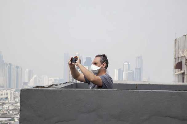 Yetişkin erkekte koruyucu maske alarak resmi cityscape smartphone cep telefonu ile çatı üzerinden - Fotoğraf, Görsel