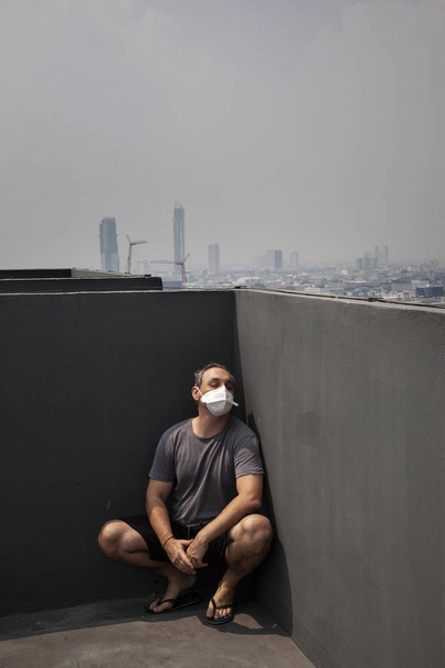 ενήλικα άνδρα που φοράει προστατευτική μάσκα κάθεται στη γωνία της στέγης με αστικό τοπίο - Φωτογραφία, εικόνα