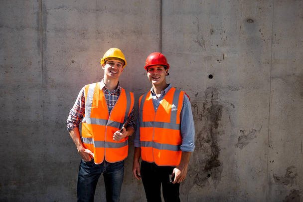 Structural engineer and architect dressed in orange work vests and  helmets stand on a concrete wall background - Foto, imagen