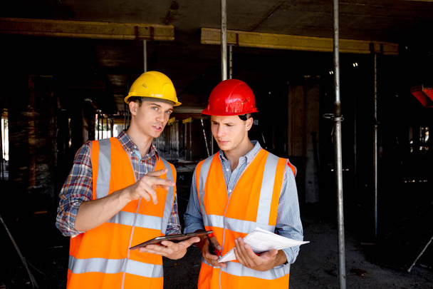 Structural engineer and architect dressed in orange work vests and  helmets use tablet and keep project documentation inside the building under construction - Zdjęcie, obraz