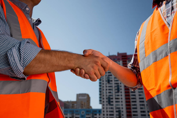 Mężczyźni ubrani w kamizelki pomarańczowy praca uścisnąć dłoń na placu budowy na tle Budynek wielokondygnacyjny - Zdjęcie, obraz
