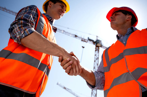 Structural engineer and architect dressed in orange work vests and  helmets shake hands on the building site near the crane - Photo, image