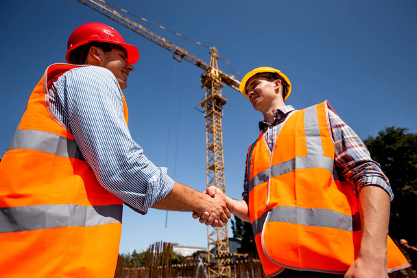 Two men dressed in orange work vests and  helmets shake hands on the building site near the crane - 写真・画像