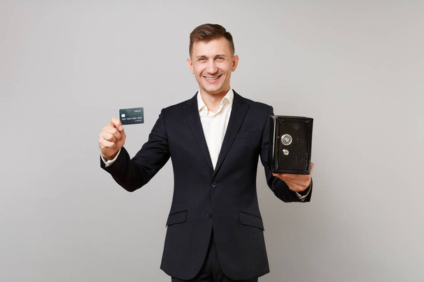Smiling young business man in classic suit holding credit bank card, metal bank safe for money accumulation isolated on grey background. Achievement career wealth business concept. Mock up copy space - Photo, Image