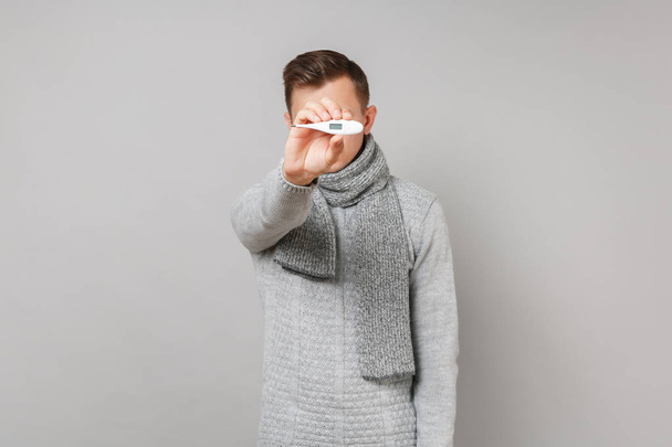 Jonge man in een grijze trui, sjaal weergegeven: thermometer op camera geïsoleerd op grijs muur achtergrond studio portret. Gezonde levensstijl, ziek zieke ziekte behandeling, koude seizoen concept. Bespotten kopie databaseruimte - Foto, afbeelding