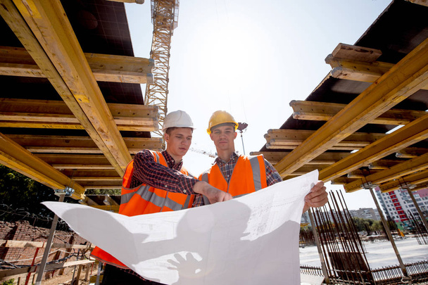 Structural engineer and architect dressed in shirts, orange work vests and helmets explore construction documentation on the building site near the wooden building constructions - Foto, Imagen