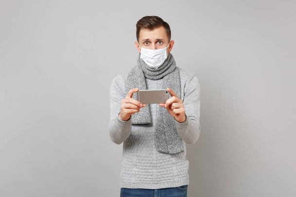 セーター マフラー滅菌マスク selfie 灰色の背景に分離されたビデオ通話携帯電話で撮影をしている若い男。健康病気病気病気治療の寒い季節の概念。コピー スペースをモックアップします。 - 写真・画像