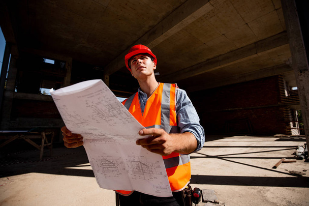 Young man dressed in shirt, orange work vest and helmet  explore construction documentation on the building site inside the building under construction - Fotografie, Obrázek