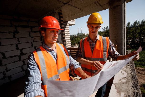 Architect  and structural engineer dressed in shirts, orange work vests and helmets explore construction documentation on the building site inside the building under construction - Φωτογραφία, εικόνα