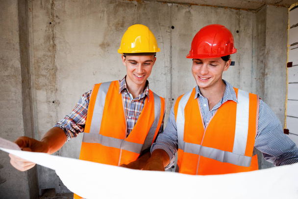 Two men dressed in orange work vests and helmets explores construction documentation on a concrete wall background - Foto, Bild