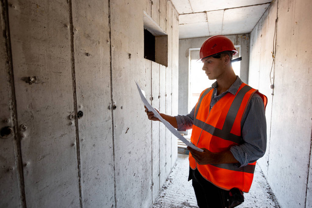 Man dressed in orange work vest and helmet explores construction documentation on a inside the building under construction - Zdjęcie, obraz