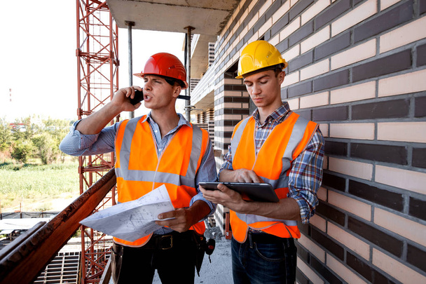 Structural engineer and architect dressed in orange work vests and  helmets discuss the construction process, use a phone and tablet on the balcony of building under construction - Foto, Bild