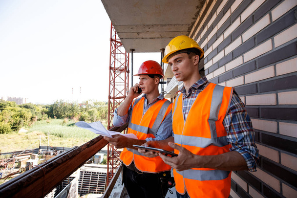 Structural engineer and architect dressed in orange work vests and  helmets discuss the construction process, use a phone and tablet on the balcony of building under construction - Foto, immagini