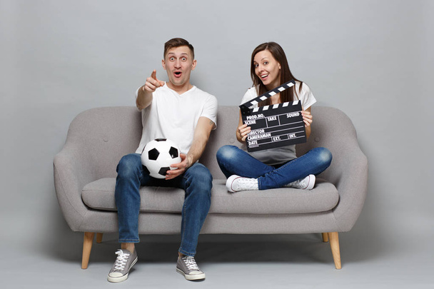 びっくりカップル女性男性サッカーファンはサッカー ボールの指を指す、灰色の背景に分離されたカチンコを作る古典的な黒いフィルムを保持をサポートの好きなチームを応援します。スポーツ家族概念 - 写真・画像